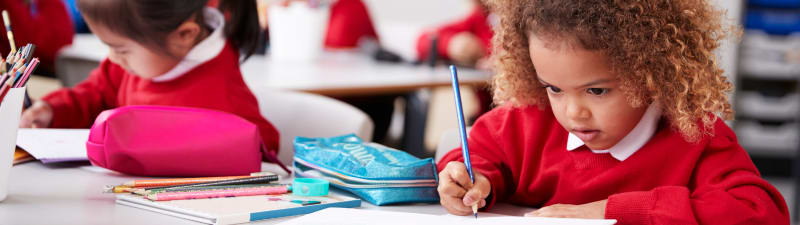 Image of a school boy wearing a red school jumper. He is sitting at a desk and writing with a pencil.